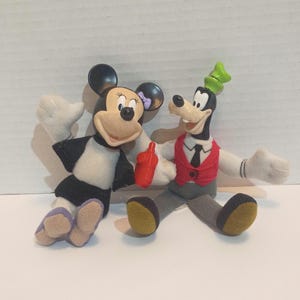 Op de afbeelding: Twee pluche Disney-personagepoppen, Minnie Mouse en Goofy, worden afgebeeld. Minnie Mouse draagt een zwart-witte jurk met een paarse strik. Goofy draagt een rood vest, een zwarte stropdas en een groene hoed. Beide hebben witte handschoenen en lachen.