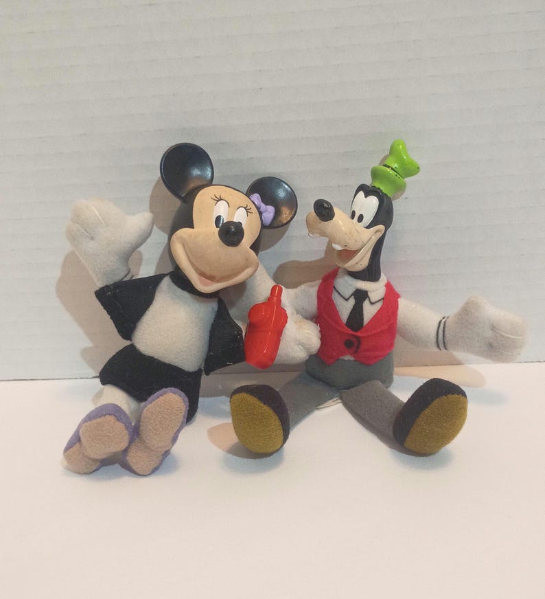 Op de afbeelding: Twee pluche Disney-personagepoppen, Minnie Mouse en Goofy, worden afgebeeld. Minnie Mouse draagt een zwart-witte jurk met een paarse strik. Goofy draagt een rood vest, een zwarte stropdas en een groene hoed. Beide hebben witte handschoenen en lachen.