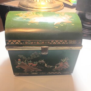 May include: A vintage green metal box with a curved top and a clasp closure. The box is decorated with gold and brown Asian-inspired landscape designs. The box has a decorative border around the top and bottom.