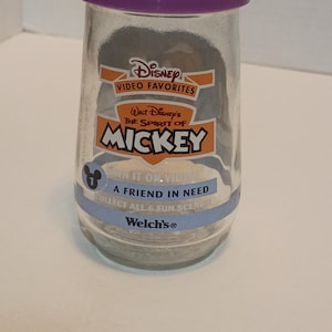 Puede incluir: Un tarro de cristal transparente con tapa morada que presenta "Video Favorites" de Disney con el texto "The Spirit of Mickey". El tarro incluye el texto "A Friend in Need" y "Collect All 6 Fun Scenes".