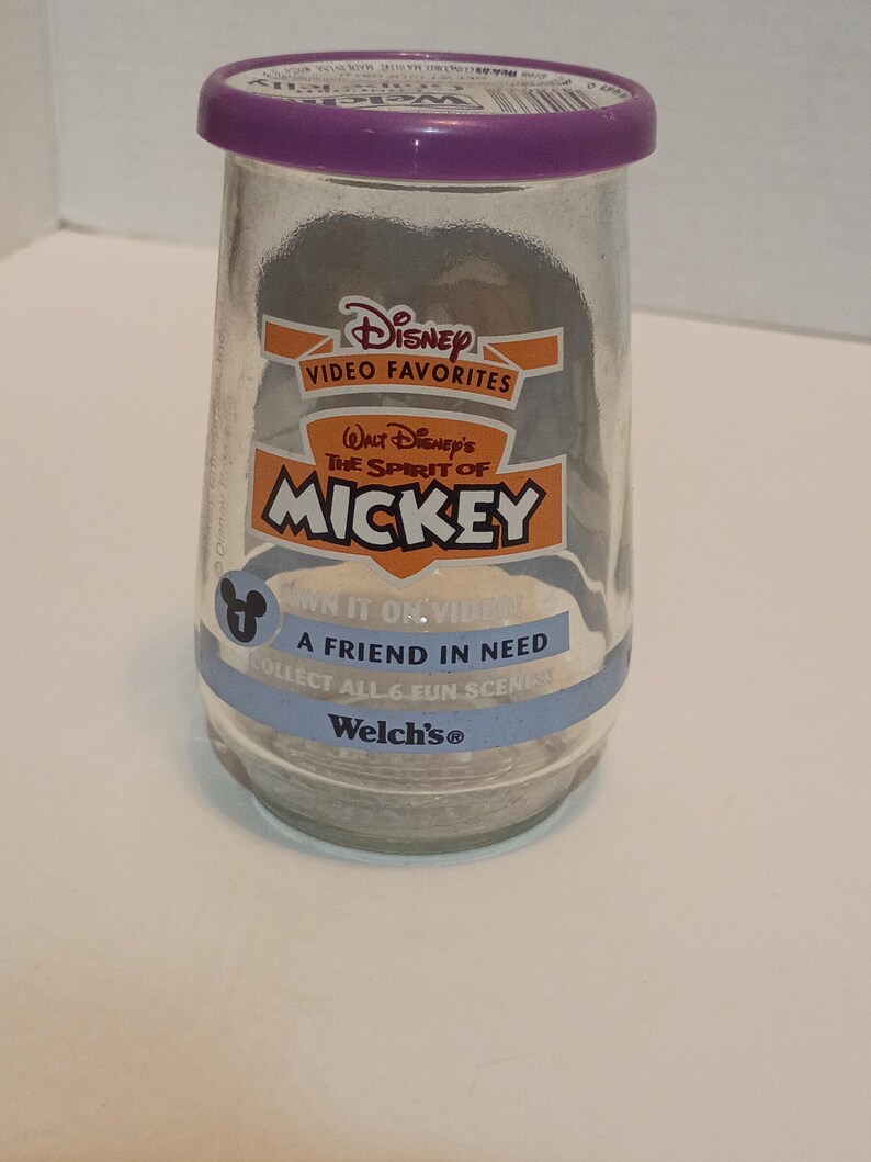 Puede incluir: Un tarro de cristal transparente con tapa morada que presenta "Video Favorites" de Disney con el texto "The Spirit of Mickey". El tarro incluye el texto "A Friend in Need" y "Collect All 6 Fun Scenes".