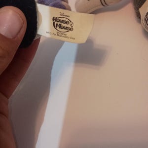 Op de afbeelding: Een close-up van een Disney "House of Mouse" label, met de tekst "Disney's House of Mouse" en "MFG. For McDonald's Corp." Het label is bevestigd aan een pluchen speelgoed, met een zwarte handschoen en een witte handschoen met zwarte strepen zichtbaar.