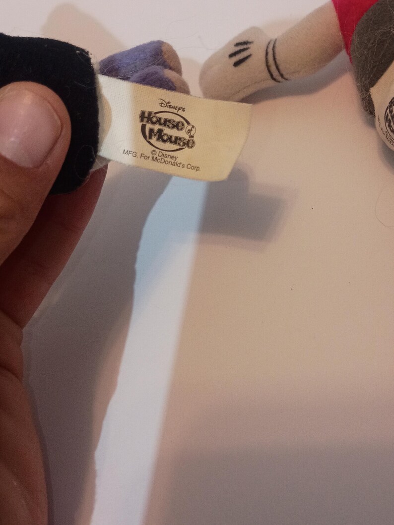 Op de afbeelding: Een close-up van een Disney "House of Mouse" label, met de tekst "Disney's House of Mouse" en "MFG. For McDonald's Corp." Het label is bevestigd aan een pluchen speelgoed, met een zwarte handschoen en een witte handschoen met zwarte strepen zichtbaar.