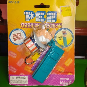Può includere: Un portachiavi torcia PEZ con lo Zio Sam, confezionato su una scheda gialla e rossa. Il portachiavi ha un corpo torcia blu e un distributore PEZ dello Zio Sam rosso, bianco e blu. La confezione include il testo "REALLY WORKS!" e "Batterie incluse."
