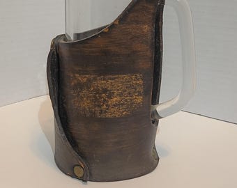 vintage 1984 clear glass beer stein featuring a brown leather sleeve with button snaps. It is approximately 6.5 inches tall with a 12oz