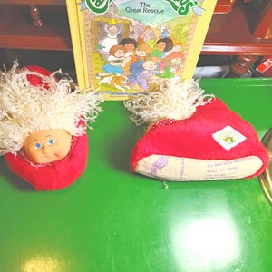 May include: Two Cabbage Patch Kids doll heads in red fabric with blonde hair, and a yellow book titled "Cabbage Patch Kids The Great Rescue" on a green surface. The doll heads have blue eyes and are in a red fabric. The book has a colorful illustration.
