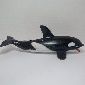 May include: A black and white orca whale figurine featuring a dorsal fin, side flippers, and a tail fluke. The whale has a white patch near the eye and a white underside, with a small open mouth showing a hint of red detail on the interior.