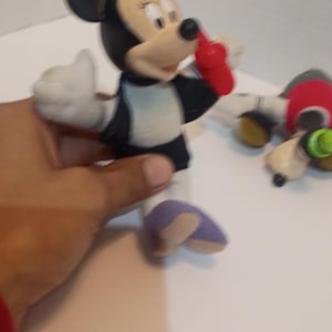 Op de afbeelding: Een Minnie Mouse-beeldje, met een zwarte kop en oren, een wit gezicht en een rode strik. Het figuur draagt een zwart-witte outfit met paarse schoenen. Een hand houdt het beeldje vast.