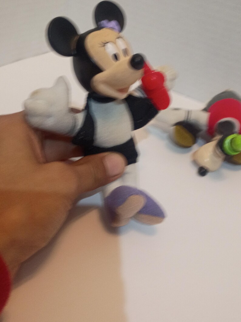 Op de afbeelding: Een Minnie Mouse-beeldje, met een zwarte kop en oren, een wit gezicht en een rode strik. Het figuur draagt een zwart-witte outfit met paarse schoenen. Een hand houdt het beeldje vast.