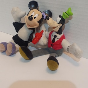 Op de afbeelding: Twee pluche Disney-personagepoppen, Minnie Mouse en Goofy, worden getoond. Minnie Mouse draagt een zwart-witte outfit met een rood accessoire. Goofy draagt een rood vest en een groene hoed. Beide poppen hebben grijze armen en benen.