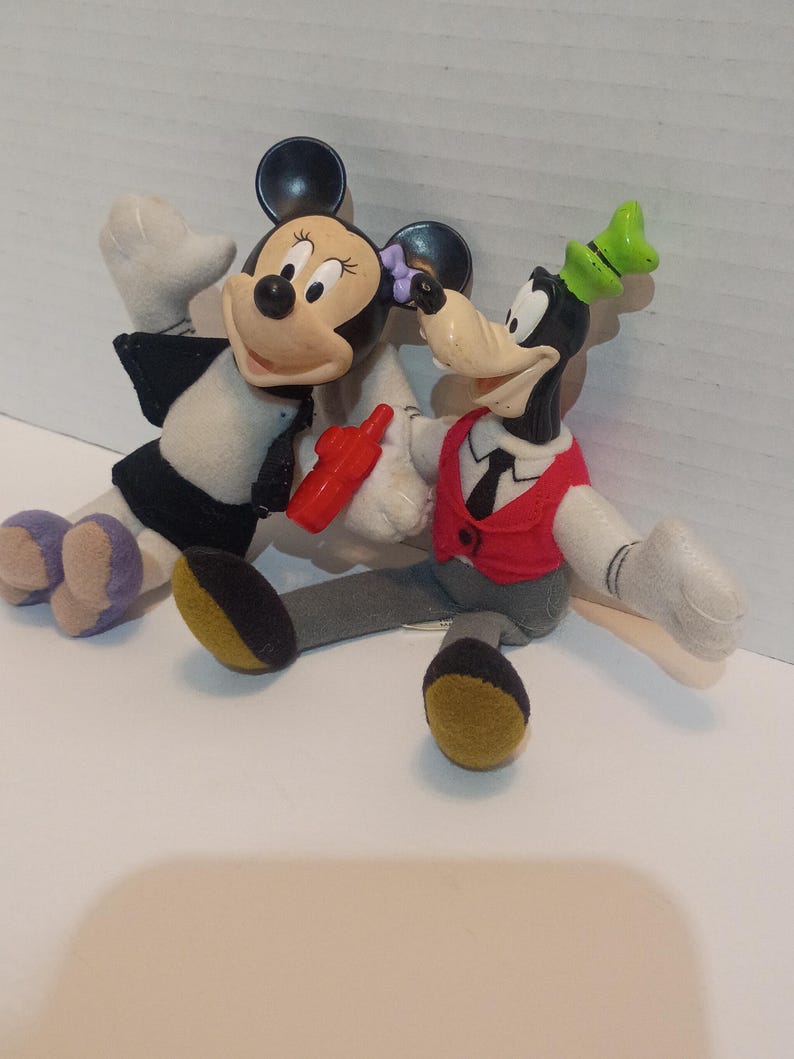 Op de afbeelding: Twee pluche Disney-personagepoppen, Minnie Mouse en Goofy, worden getoond. Minnie Mouse draagt een zwart-witte outfit met een rood accessoire. Goofy draagt een rood vest en een groene hoed. Beide poppen hebben grijze armen en benen.