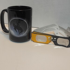 May include: Black ceramic mug featuring a silhouette of a cowboy on a bucking bronco against a moonlit background. Next to the mug are solar eclipse glasses with the date Aug. 21, 2017 printed on them.