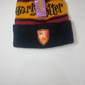 May include: A knit beanie featuring horizontal stripes in maroon and gold with the text Harry Potter in black lettering. The cuff is solid black and displays a shield-shaped patch with a lion emblem and the word Gryffindor in the center.