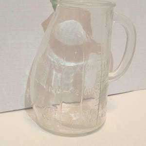 May include: A clear glass measuring pitcher with a handle and a pouring spout. The exterior features embossed measurement markings in cups, including 1/2 cup, 1 cup, 1 1/2 cups, and 2 cups, along with corresponding pint and fluid ounce indicators.