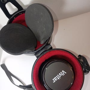 May include: Black Vivitar lens cap inside a black, round case with a red interior. The case has a black strap and two black foam inserts. The case is open, revealing the lens cap and foam padding.