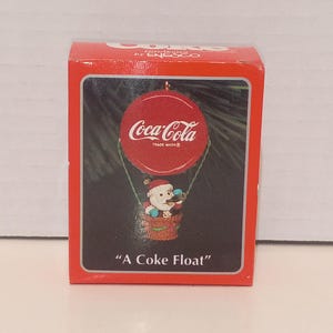 May include: A red rectangular box featuring a Coca-Cola ornament titled A Coke Float. The ornament depicts a Santa Claus figure in a basket suspended from a red bottle cap with the Coca-Cola logo. The box has white text and branding.