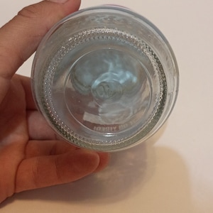 Puede incluir: Botella de vidrio transparente con base texturizada y forma redondeada. La botella es sostenida por una mano, mostrando su transparencia. La base tiene un dise&ntilde;o circular.