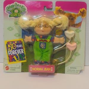 May include: A Cabbage Patch Kids doll named Norma Jean, packaged in a pink and green blister pack. The doll has blonde pigtails, glasses, and wears a green jumpsuit with blue overalls. The packaging includes "Toys R Us 50 Years Forever Fun" text.