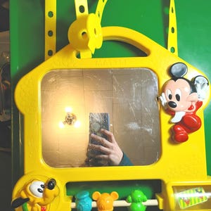May include: Yellow plastic toy with a mirror, featuring Mickey Mouse and Pluto characters. The toy has a handle and a strap, with colorful spinning elements and small figures. The word "Mattel" is visible on the toy.