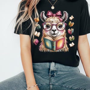 May include: Black t-shirt featuring a whimsical illustration of a llama wearing glasses and a pink bow, reading a book. The llama is surrounded by floating books in various colors. A fun and unique design for book lovers.