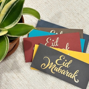 Puede incluir: Un conjunto de tarjetas rectangulares de colores con la inscripción dorada «Eid Mubarak» y «Eid». Las tarjetas son de color gris, rojo, turquesa, marrón y amarillo. En el fondo, una planta en maceta con hojas verdes y amarillas.