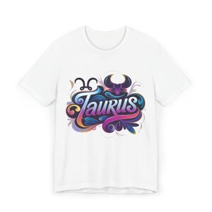 Peut inclure: T-shirt blanc avec un motif coloré du signe astrologique du Taureau. Le motif comprend le mot "Taurus" dans une police stylisée avec des teintes bleues, violettes et roses, ainsi qu'un symbole de taureau et des volutes décoratives.