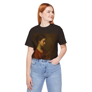 Peut inclure: T-shirt noir avec une illustration vintage d'une femme et d'un scorpion. L'œuvre d'art utilise des tons marron, rouge et or, avec le symbole du zodiaque Scorpion. Le t-shirt est en matière douce et a des manches courtes.
