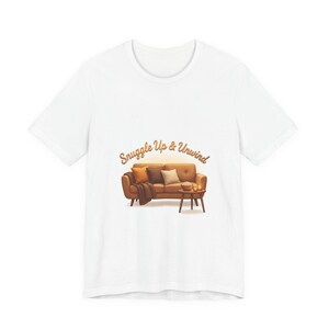 May include: White t-shirt featuring a graphic of a brown sofa with pillows, a blanket, and a side table with a bowl of popcorn. The text "Snuggle Up & Unwind" is arched above the sofa in a warm brown color.
