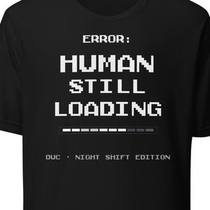 Op de afbeelding: Zwart t-shirt met witte pixeltekst: "ERROR: HUMAN STILL LOADING" boven een laadbalk. De tekst "DUC - NIGHT SHIFT EDITION" staat eronder.