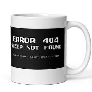 Può includere: Tazza in ceramica bianca con una fascia nera con la scritta "ERROR 404 SLEEP NOT FOUND" in bianco, con font pixelato. Sotto c'è la scritta "DIAL-UP CLUB NIGHT SHIFT EDITION". La tazza ha un manico curvo.