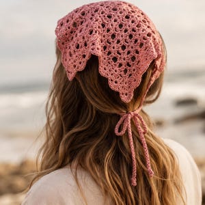 May include: A dusty rose crochet headscarf featuring an intricate openwork pattern with a scalloped edge. The triangular head covering is secured at the back of the head with two thin, matching crocheted ties that are knotted into a bow.