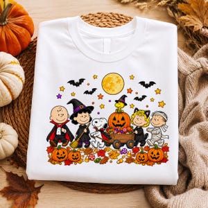 May include: A white t-shirt featuring a colorful graphic of Peanuts characters in Halloween costumes, including a vampire, a witch, a mummy, and a cat, with Snoopy pulling a wagon of pumpkins under a full moon, bats, and autumn leaves.