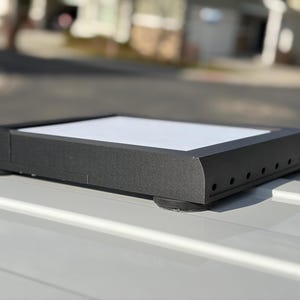 Könnte beinhalten: Ein rechteckiges schwarzes Objekt mit einer weißen Oberfläche, das auf einer weißen Oberfläche ruht. Das schwarze Objekt hat abgerundete Ecken und kleine kreisförmige Löcher entlang einer Seite. Es wird von kleinen, runden, schwarzen Füßen angehoben.