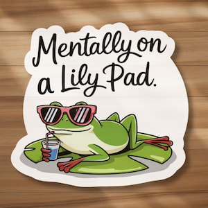May include: A whimsical sign featuring a cartoon frog wearing sunglasses and holding a drink, lounging on a lily pad. The sign has the text "Mentally on a Lily Pad" in black script, set against a white background with a wooden texture.