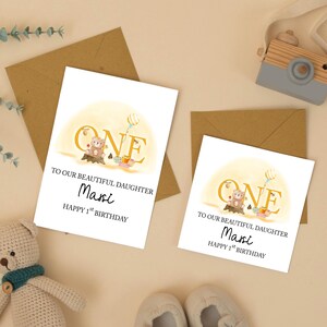May include: Two white greeting cards with the text "ONE" and "TO OUR BEAUTIFUL DAUGHTER Maxi HAPPY 1ST BIRTHDAY". The cards feature a teddy bear illustration and are placed on a light brown surface with matching envelopes.