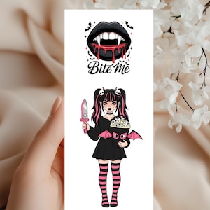 Può includere: Un'illustrazione digitale con labbra nere con sangue che cola e la scritta "Bite Me". Sotto, una figura di cartone animato di una ragazza con capelli rosa e neri, che tiene in mano un coltello e un contenitore a forma di pipistrello. Indossa un vestito nero e calzini a righe.