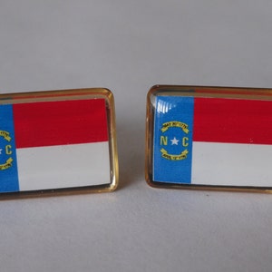 K&ouml;nnte beinhalten: Ein Paar goldfarbene Manschettenkn&ouml;pfe mit der Flagge des Bundesstaates North Carolina. Die Flagge ist blau, wei&szlig; und rot mit einem gelben Stern und dem Text "MAY 20, 1775 APRIL 12, 1776" und "N.C."