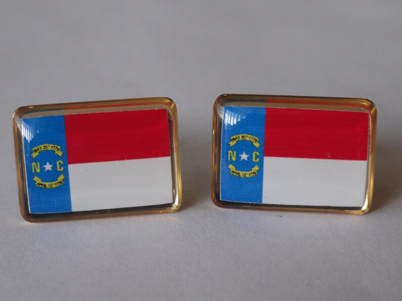 K&ouml;nnte beinhalten: Ein Paar goldfarbene Manschettenkn&ouml;pfe mit der Flagge des Bundesstaates North Carolina. Die Flagge ist blau, wei&szlig; und rot mit einem gelben Stern und dem Text "MAY 20, 1775 APRIL 12, 1776" und "N.C."