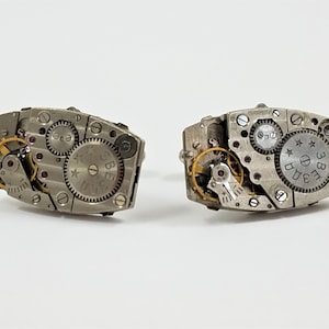 May include: A pair of silver cufflinks with a vintage watch movement design. The cufflinks feature exposed gears and a star-shaped dial with the text "3 BE 3B" on it.