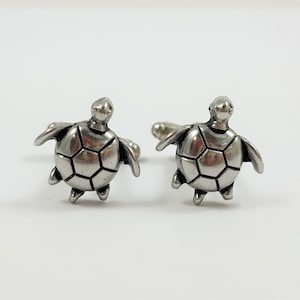 May include: A pair of silver cufflinks featuring a turtle design. The turtles are facing forward with their heads up and their legs outstretched.