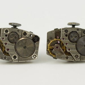 Puede incluir: Un par de gemelos de plata con mecanismos de relojería expuestos. Los gemelos son de forma ovalada y tienen un acabado plateado. La relojería es visible a través de la parte posterior abierta de los gemelos.