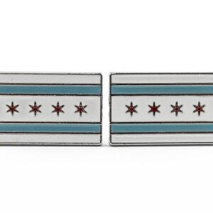 Op de afbeelding: Een paar zilveren manchetknopen met de vlag van Chicago. De vlag is wit met twee blauwe horizontale strepen en zes rode sterren.