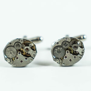 Puede incluir: Un par de gemelos de plata con mecanismos de relojería expuestos. Los gemelos son de forma ovalada y tienen un acabado plateado.