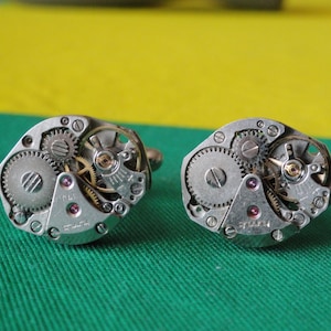 Puede incluir: Un par de gemelos de plata con intrincados diseños de relojería. Los engranajes son visibles y los gemelos tienen un acabado pulido.