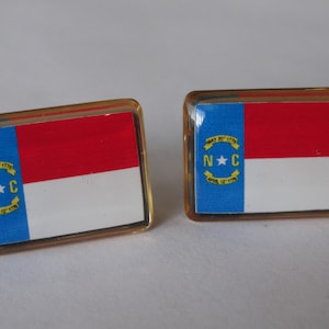 K&ouml;nnte beinhalten: Zwei goldfarbene Manschettenkn&ouml;pfe mit der Flagge des Bundesstaates North Carolina. Die Flagge ist blau, wei&szlig; und rot mit einem gelben Stern und den Buchstaben "N.C." in einem gelben Kreis.