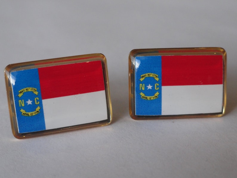 K&ouml;nnte beinhalten: Zwei goldfarbene Manschettenkn&ouml;pfe mit der Flagge des Bundesstaates North Carolina. Die Flagge ist blau, wei&szlig; und rot mit einem gelben Stern und den Buchstaben "N.C." in einem gelben Kreis.