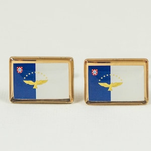 May include: A pair of gold cufflinks featuring the flag of the Azores, a Portuguese autonomous region. The flag is blue with a white field on the right side, a gold eagle, and a small red shield with a white cross.