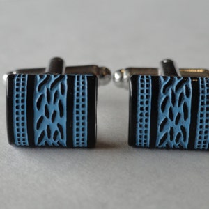May include: A pair of cufflinks with a black and blue design. The cufflinks have a silver metal base and a rectangular blue and black enamel top. The design features a woven pattern in blue on a black background.
