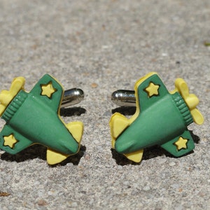 Puede incluir: Un par de gemelos novedosos con forma de aviones verdes y amarillos. Cada gemelo presenta un cuerpo verde con alas y hélice amarillas, y detalles de estrellas amarillas. Los gemelos tienen postes plateados.