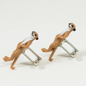 May include: A pair of cufflinks featuring a realistic meerkat figurine on each cufflink. The meerkats are brown and white and are posed in a standing position. The cufflinks are silver.